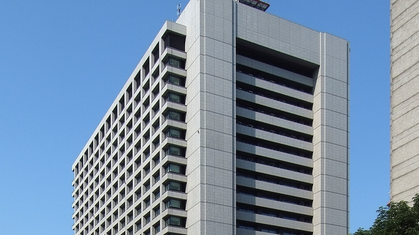 A grey government office tower in Tokyo photographed from below against a clear blue sky. The nearer wing is a dense grid of small square recessed windows running the full height of the elevation, and the taller wing beside it is faced in smooth pale stone panels with a deep vertical recess of stacked balcony bands down its centre. A dark window-cleaning gondola rail with a red edge projects from the parapet at the top. A second concrete tower stands at the right edge of the frame and the crown of a green tree enters at the bottom right corner. No people, vehicles or signs are visible.