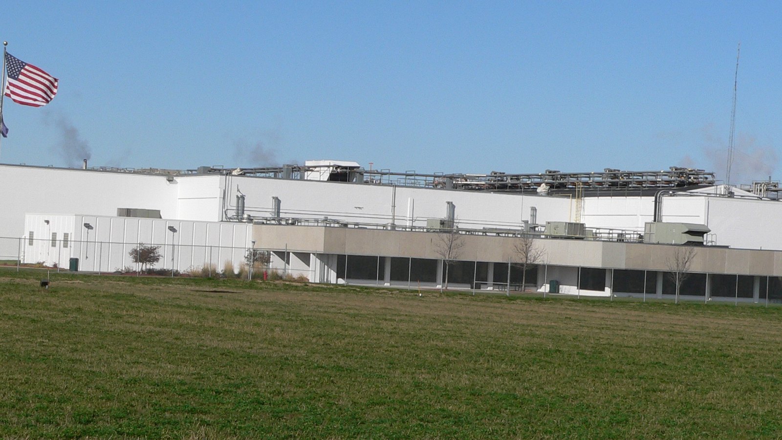 A long white food processing plant seen across a mown green field from behind a chain-link fence, with pipework and exhaust stacks running the length of the roof and an American flag on a pole at the left edge.