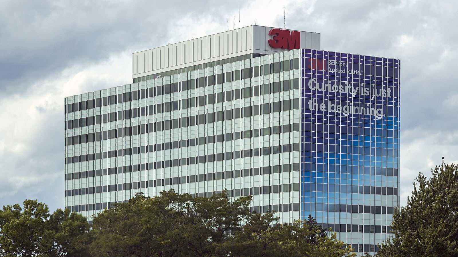 The 3M headquarters tower in Maplewood, Minnesota, a pale glass office block with a red 3M logo on its rooftop and a blue facade panel carrying the company slogan, rising above a line of trees under grey cloud.