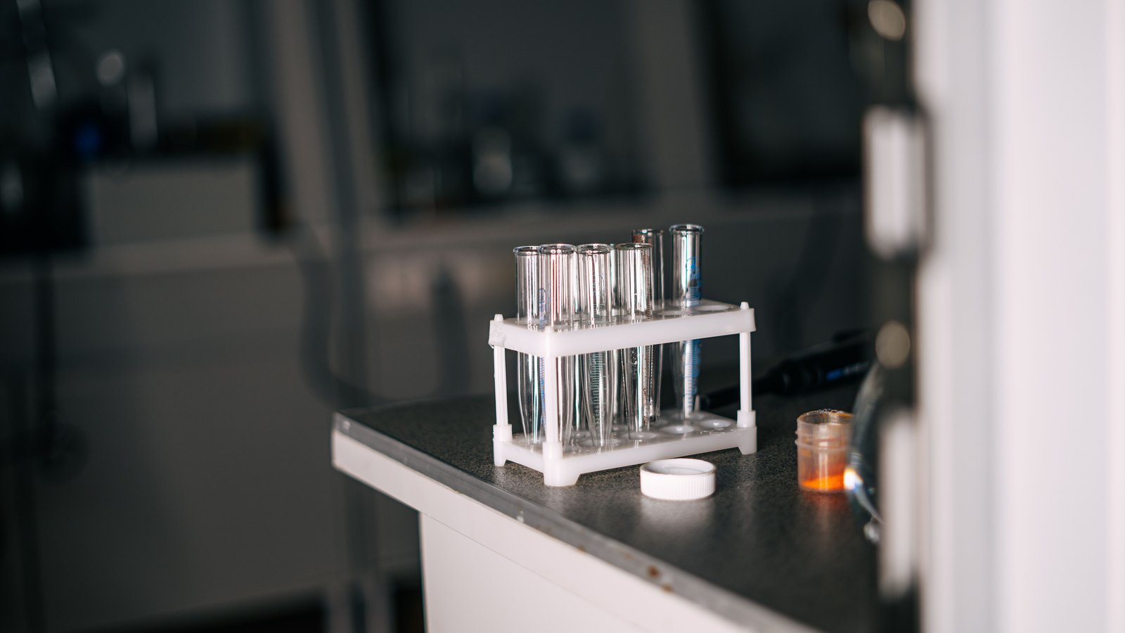 A white rack of clear glass test tubes on a grey laboratory bench, beside a small amber sample jar and a white cap, with the lab blurred behind.