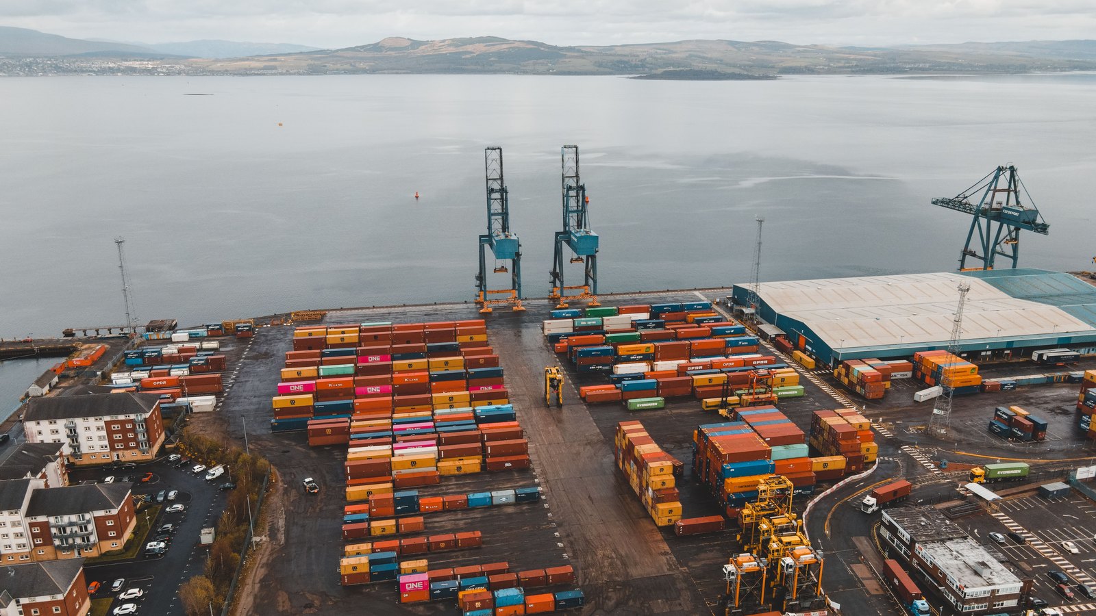 An aerial view of a container terminal beside a broad harbor, with rows of colorful containers and three dockside cranes.