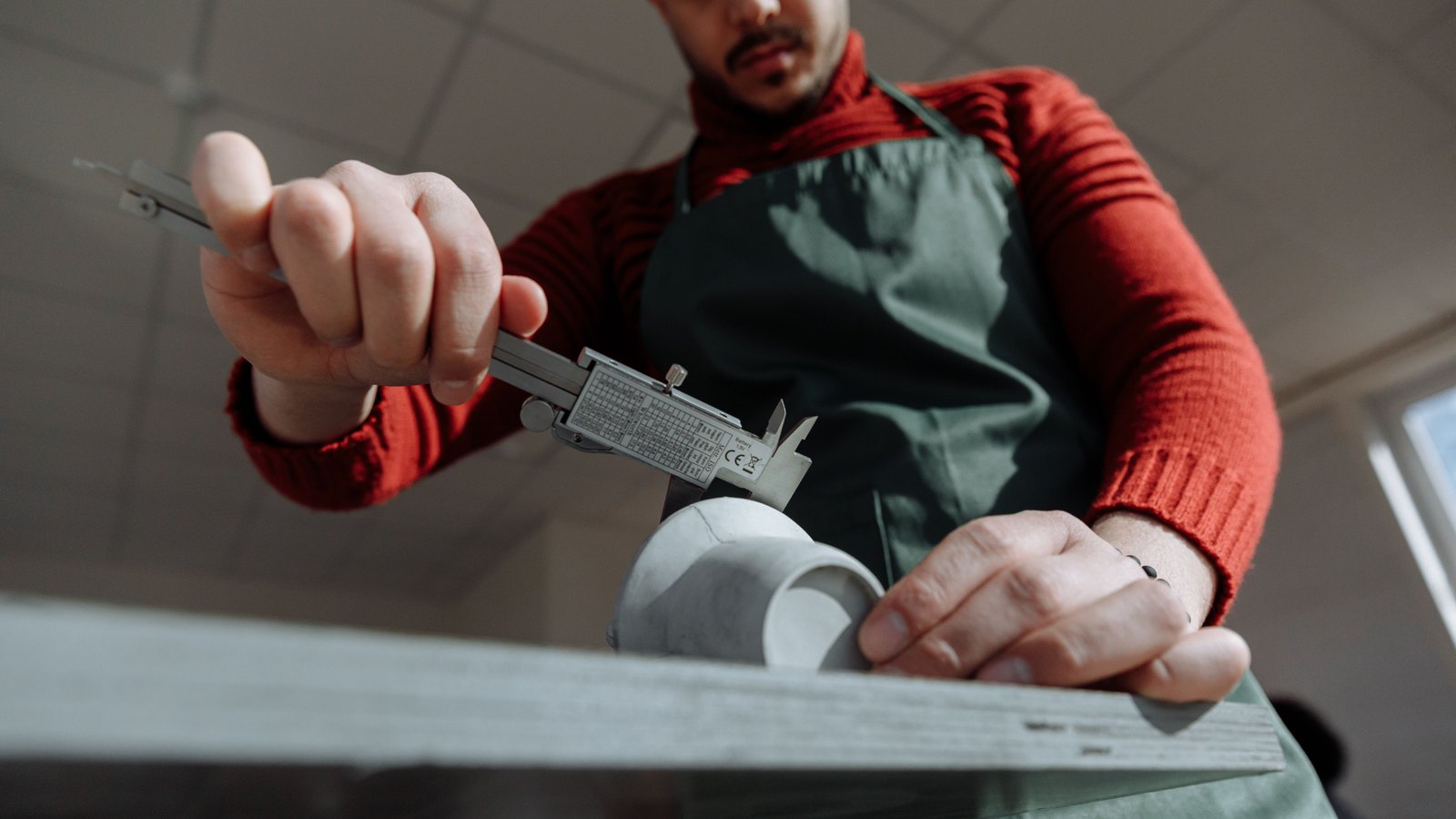A person in a work apron measures the edge of a pale component with a steel vernier caliper on a workshop bench.