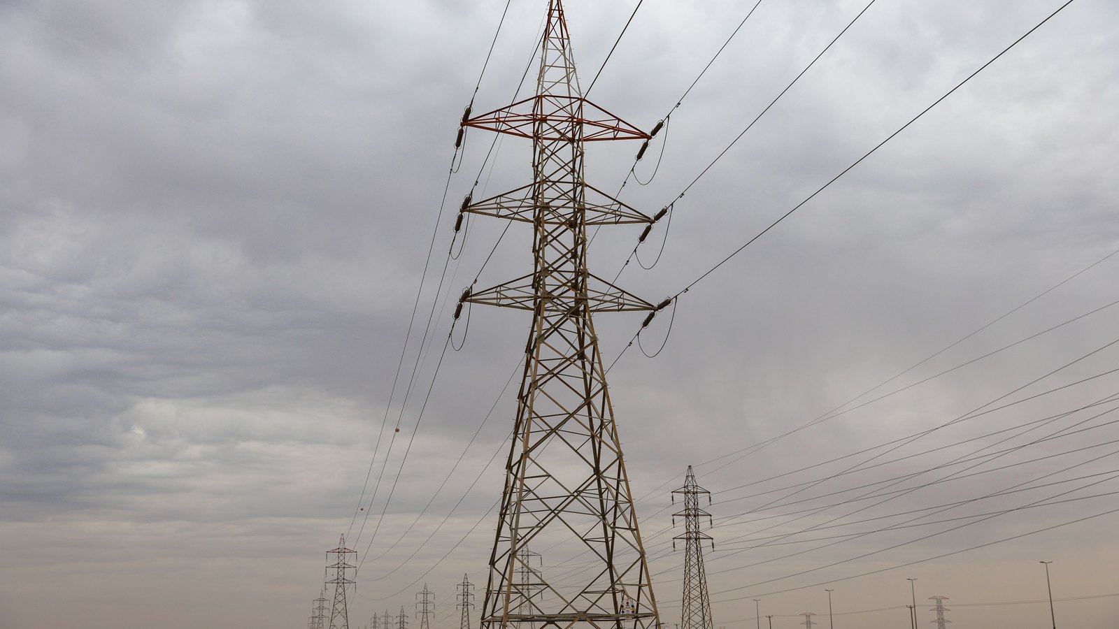 A steel lattice electricity transmission pylon photographed from below against a flat grey overcast, its conductors fanning out to both sides on strings of insulators, with further pylons receding into haze along the line behind it. There are no people, vehicles, buildings or signs in the frame.