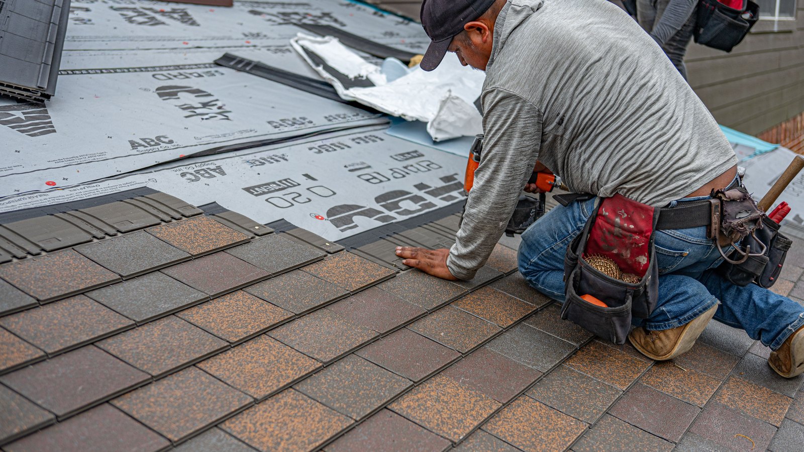 A roofer kneeling to lay rows of tan and brown asphalt shingles over roofing underlayment on a residential roof.
