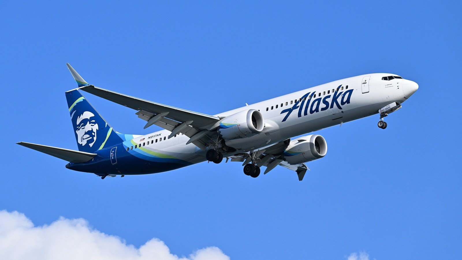 An Alaska Airlines Boeing 737-9 MAX in the carrier's white and blue livery, with the carrier portrait on the tail, flying on approach against a clear blue sky with its landing gear down and a cloud at the lower left.