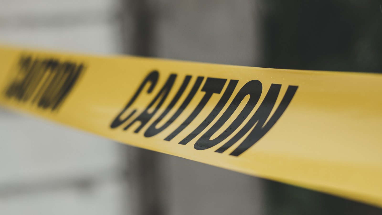 A close-up of yellow barrier tape printed with the word CAUTION in black capitals, stretched diagonally across the frame, with a pale wall and dark greenery blurred out of focus behind it, and no people, vehicles, number plates, signs, agency markings or place names anywhere in the frame. A generic illustration of a cordoned scene, not a photograph of this event.