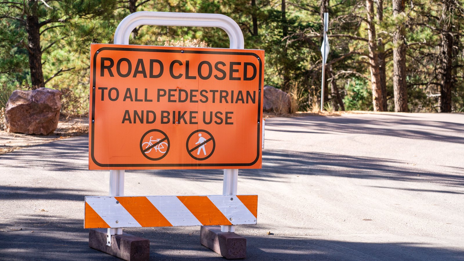 An orange road-closure sign and striped barricade on a paved route among trees, not a photograph of this event.