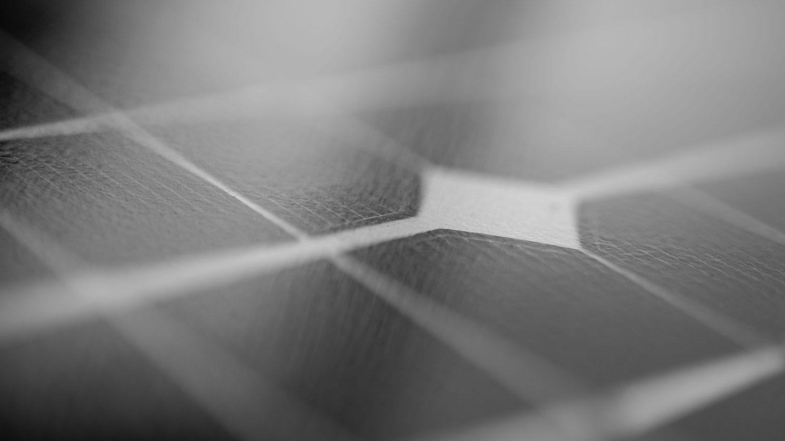 A monochrome close photograph of the face of a photovoltaic cell, the pale silver busbars crossing the darker cell squares in a shallow grid that runs out of focus towards the top of the frame.