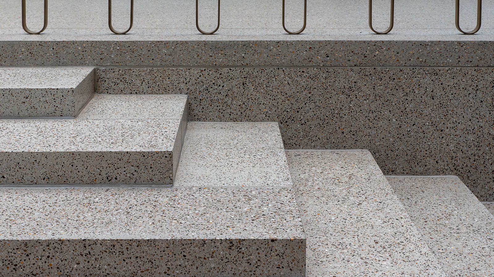 Wide terrazzo steps in speckled grey and cream aggregate rising in shallow offset tiers, with slim metal railing loops running along the top edge.