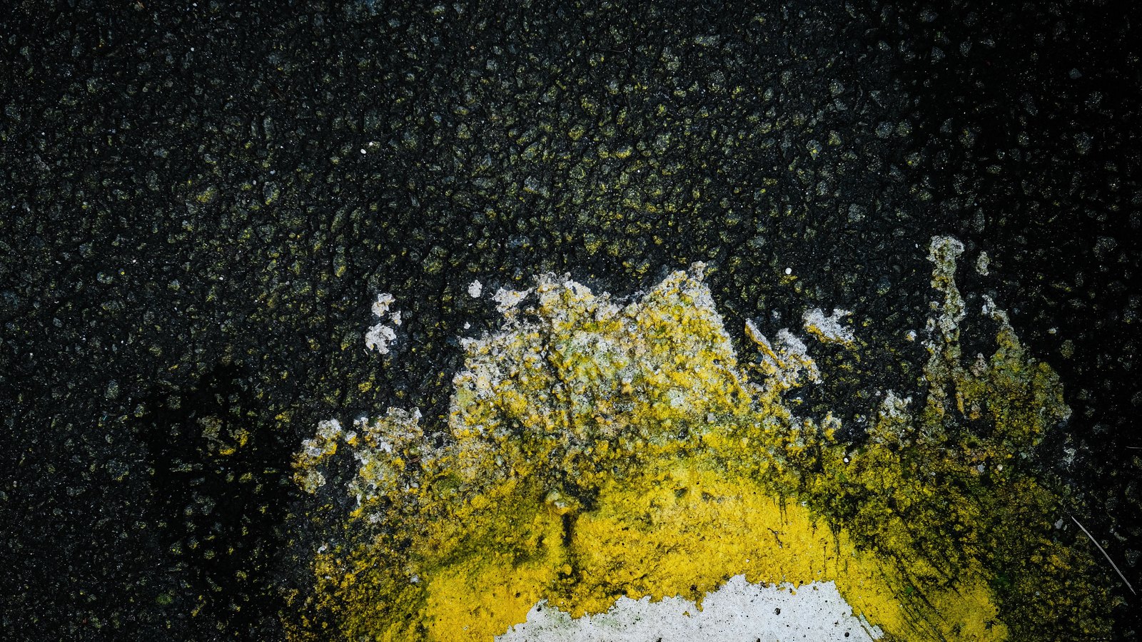 A tight overhead macro of coarse dark asphalt, with a worn band of yellow paint and a patch of white paint flaking across the aggregate in the lower half of the frame.