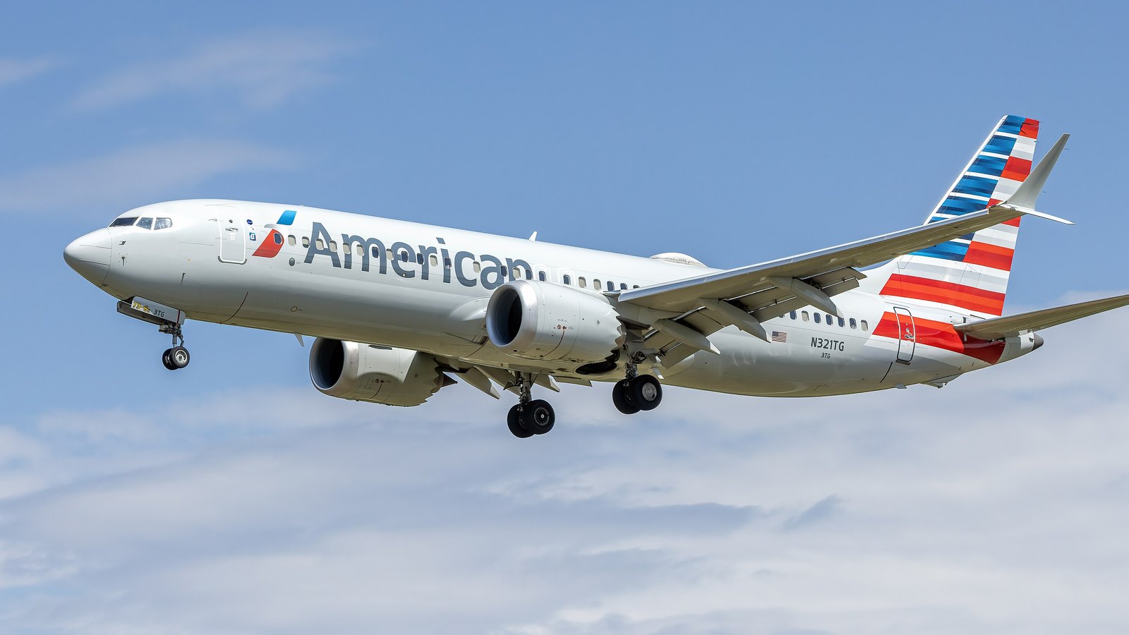 A silver American Airlines Boeing 737 MAX with red, white and blue tail stripes and its landing gear down, flying against a blue sky with thin cloud.