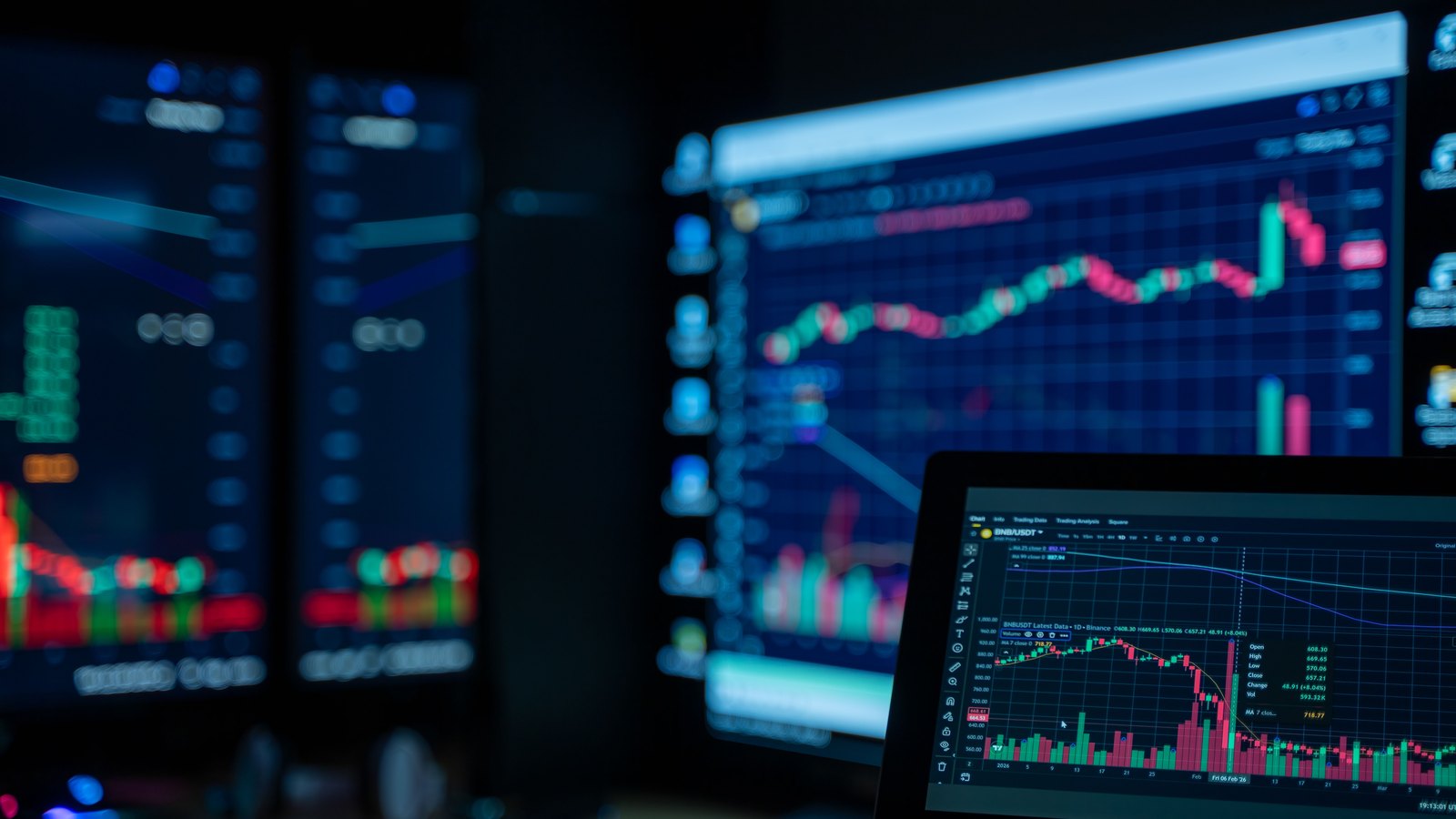 Several dark monitors and a laptop on a trading desk showing candlestick charts in red and green. No identifiable person, place or company appears. A generic illustration of financial markets.