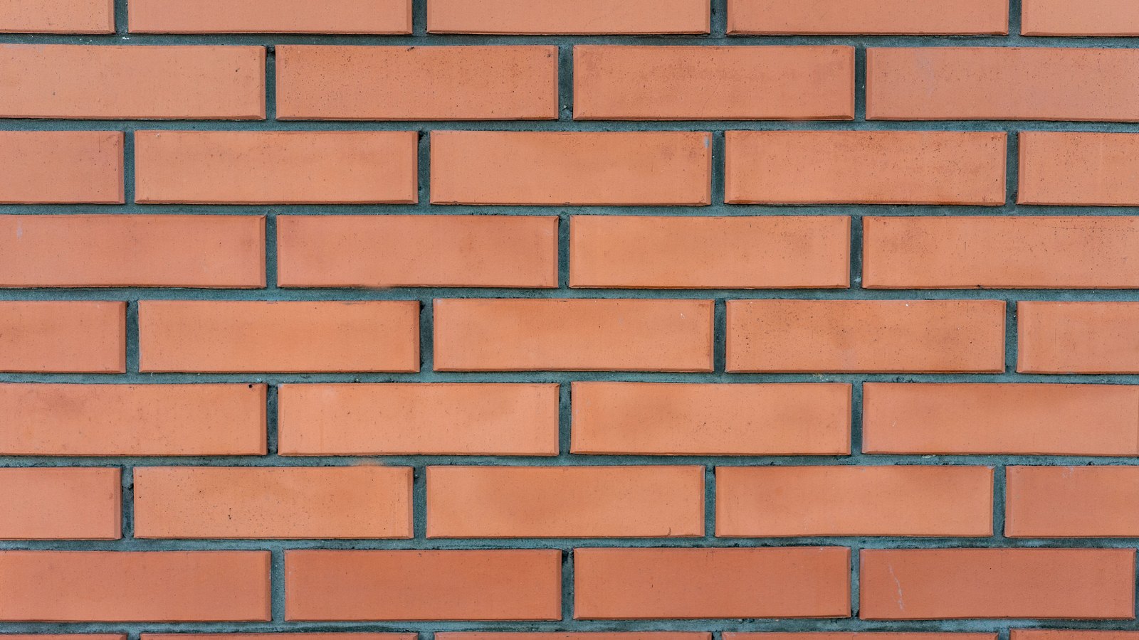 A close photograph of an orange brick wall filling the whole frame, laid in even courses of identical stretcher bricks offset by half a brick on each row, separated by narrow dark grey mortar joints, with no sky or horizon in view.