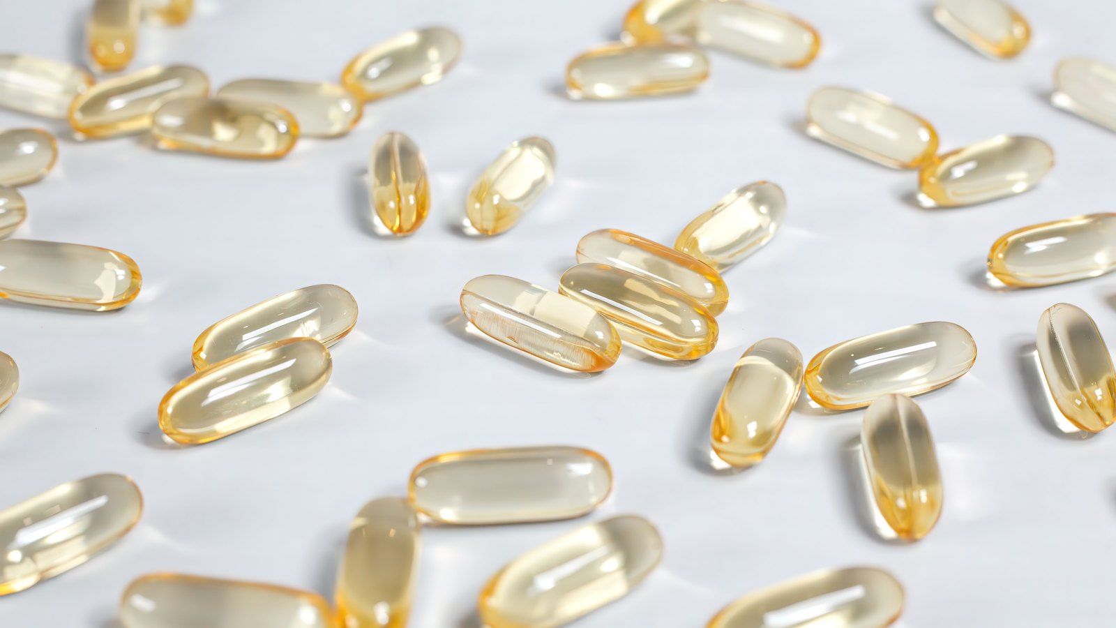Several dozen translucent golden softgel capsules scattered across a plain white surface and lit from above, with no packaging, no lettering and nobody in the frame.