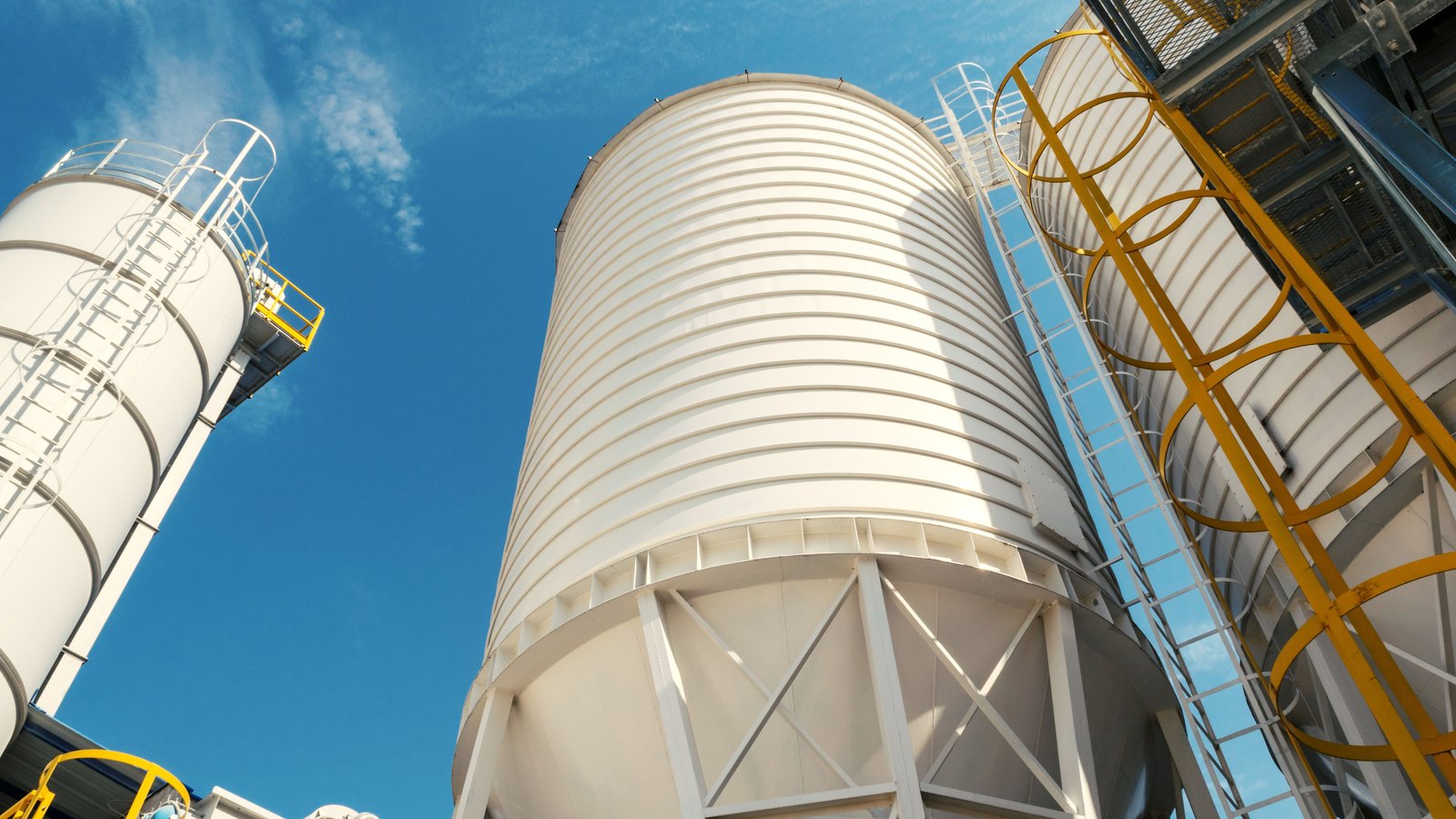 White cylindrical storage silos with yellow caged access ladders and steel walkways photographed from below against a clear blue sky.