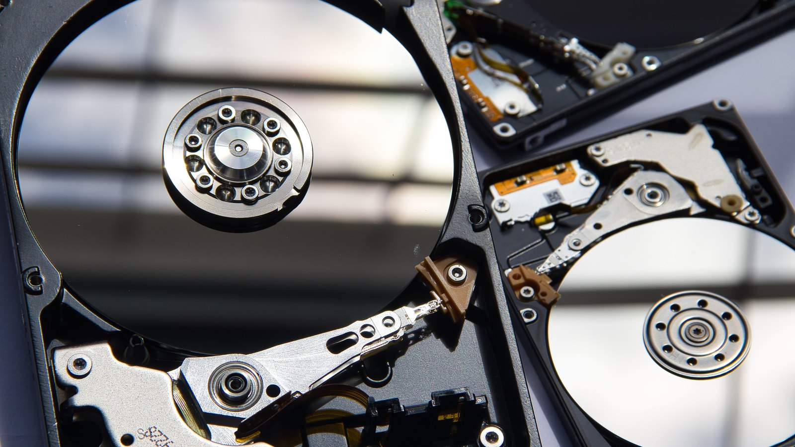 A close photograph of three opened mechanical hard drives lying on a pale surface, their mirrored platters, spindle bearings and slim actuator arms exposed, with no brand name or readable label in view.