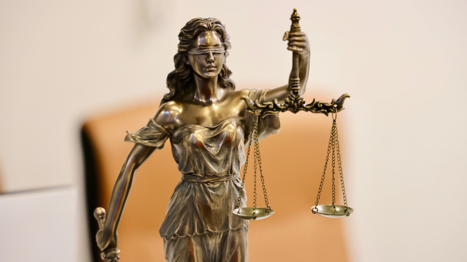 A small bronze statue of blindfolded Justice holding a set of scales that hang unevenly, against a plain blurred background. Stock photo, not a photograph of this event.