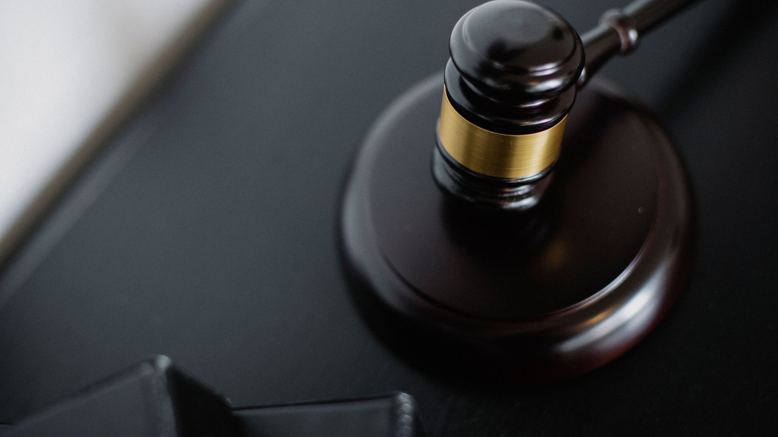 A dark polished wooden gavel resting on its round sound block on a black desk, lit from one side, with the corner of a plain black folder in the foreground and no people, faces, signage, text, logos or identifiable location anywhere in the frame. A generic illustration of a court proceeding, not a photograph of this event.