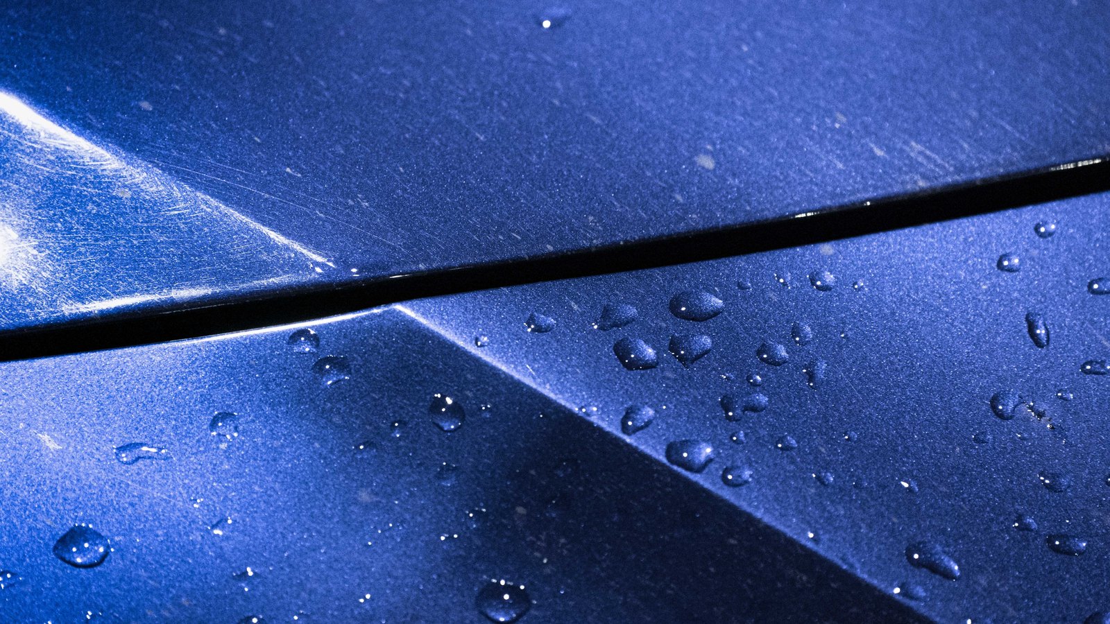 A close photograph of a deep blue metallic painted surface lit from the left, a narrow dark seam running across it from edge to edge, with beads of rainwater standing on the lower half and the metallic flake in the paint catching the light.