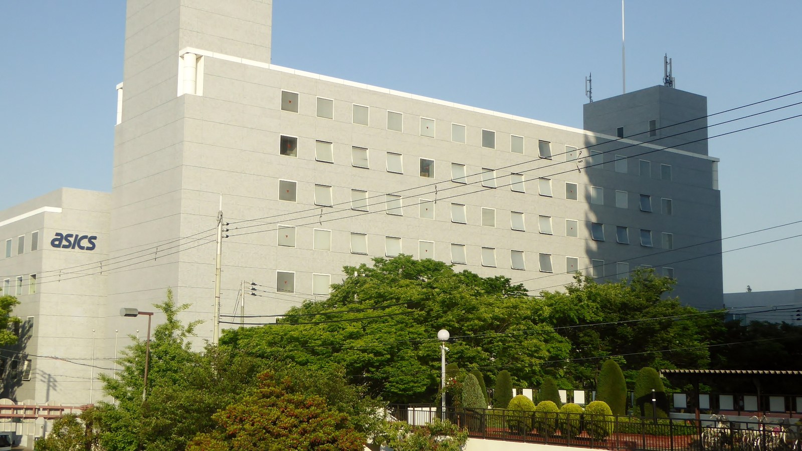 A five storey pale stone office block seen from across a road on a clear day, with a lower wing at the left carrying the ASICS wordmark in dark blue lettering. Trees, a street lamp and overhead cables cross the lower part of the frame and a taller grey building stands behind at the right. No people, vehicles, other company marks or road signs are in view.