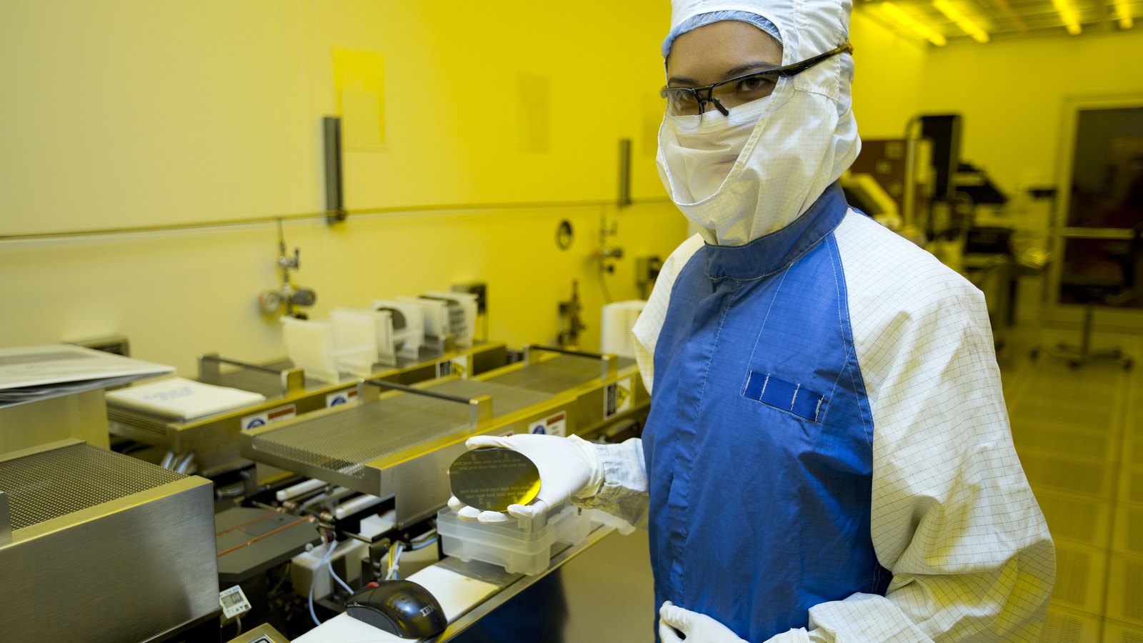 A semiconductor cleanroom worker holds a silicon wafer beside processing equipment