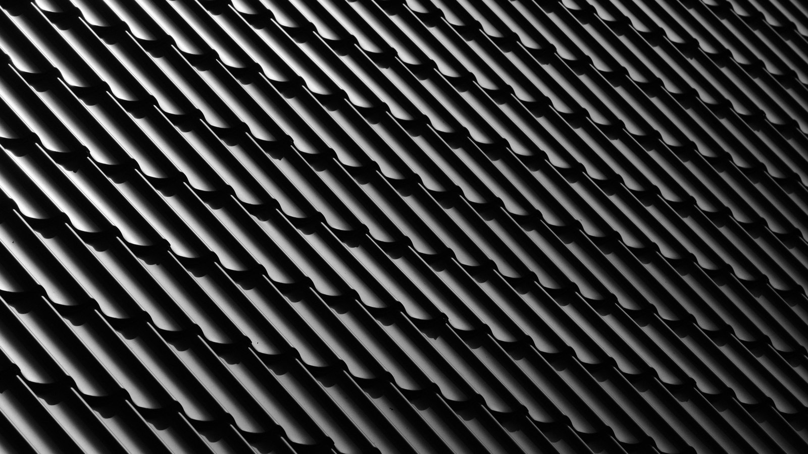 A dark profiled metal surface photographed at an oblique angle, its long parallel ribs running diagonally across the whole frame with a bright highlight along the crest of each rib and deep shadow in the channels between them. No text, brand mark, person, horizon or background is in view.