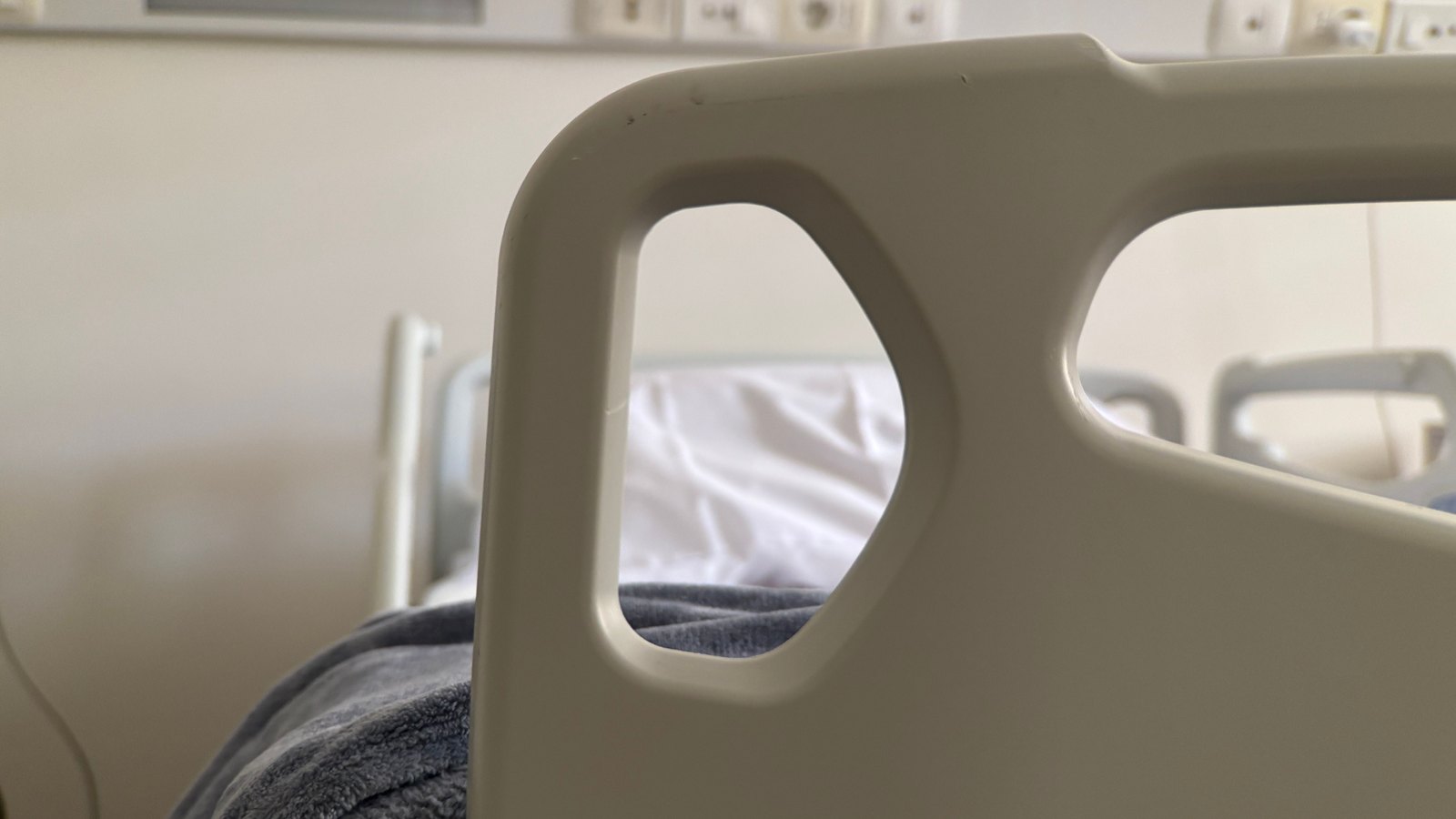 A close view of the moulded plastic side rail of a care bed, with white sheets and a folded grey blanket behind it and a second empty bed further into the room, against a plain pale wall. No people, signage or lettering appear. A generic illustration of a residential care setting, not a photograph of this event.
