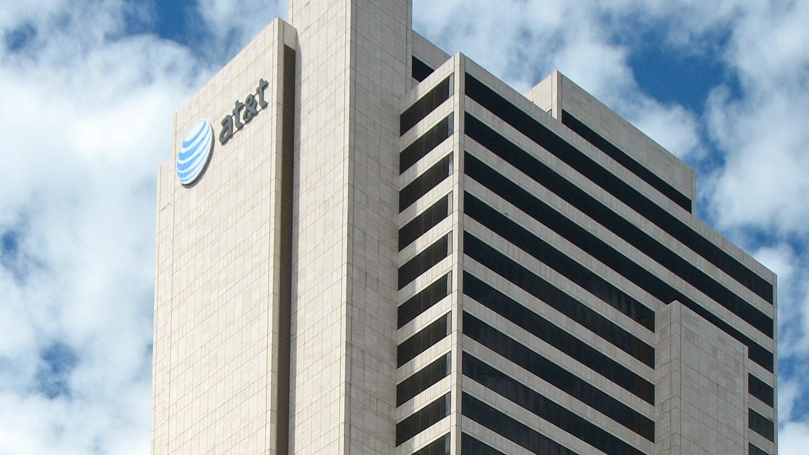 The upper storeys of a pale stone office tower in downtown Dallas carrying the AT&T globe logo and name, against a blue sky with white clouds.