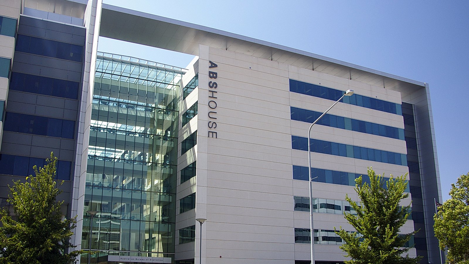 ABS House in Belconnen, Canberra, the headquarters of the Australian Bureau of Statistics: a pale stone and blue glass office block with a full height glazed atrium, ABS HOUSE lettered vertically down the facade, and the agency name on a sign above the ground floor entrance.
