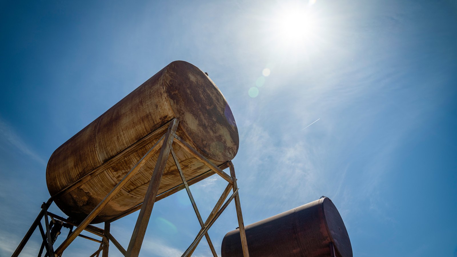 Two weathered cylindrical oil storage tanks stand on steel frames beneath a bright blue sky.