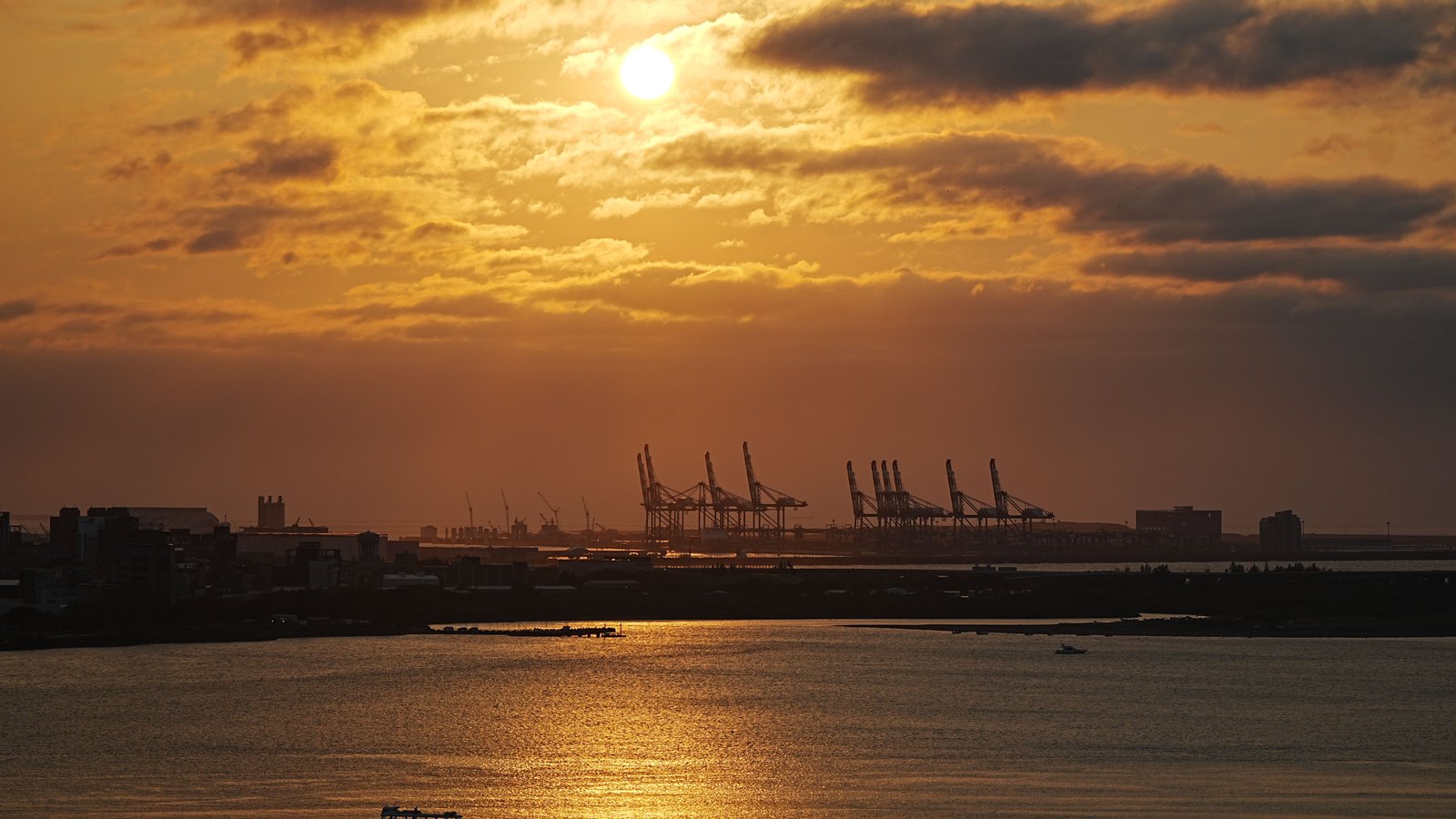 Container cranes stand in silhouette across a harbor at sunset.