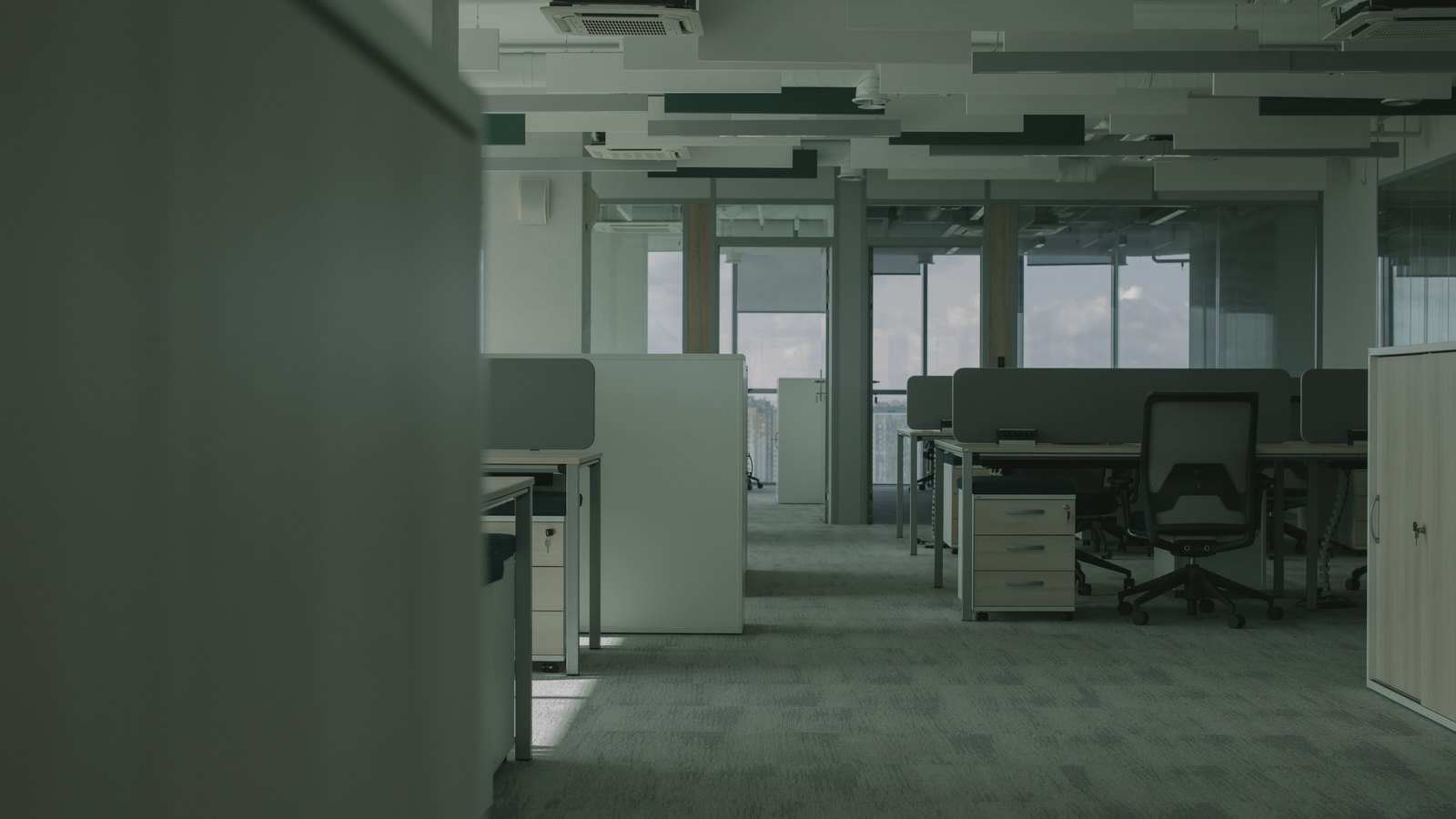 An empty open plan office floor, with rows of desks behind low privacy screens, vacant chairs, cabinets on castors and grey carpet, daylight entering through a wall of windows. An illustrative photograph, not a picture of any workplace counted in these estimates.