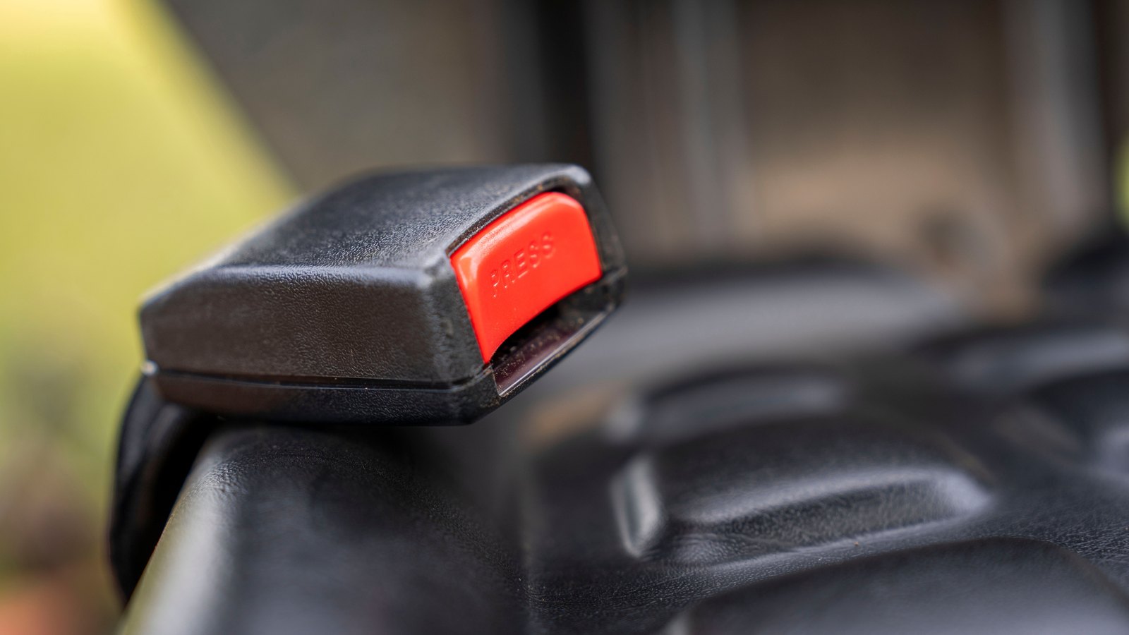 A black car seatbelt buckle with a red release button resting on a dark moulded seat, photographed close up with a soft green background.