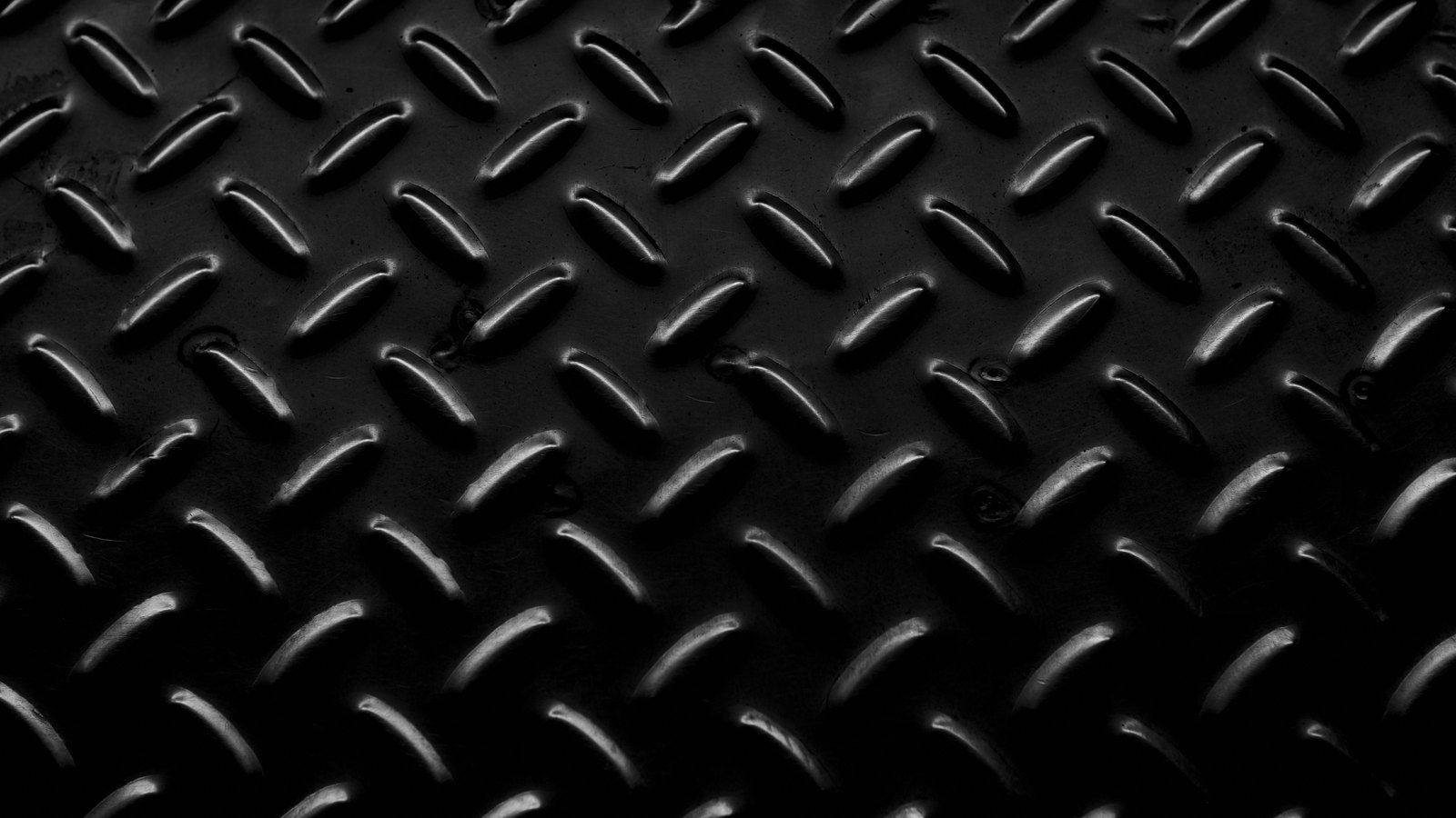 A close photograph of black steel chequer plate, rows of raised oval treads set in alternating diagonals catching a hard light along their upper edges, filling the frame and darkening towards the lower corners.