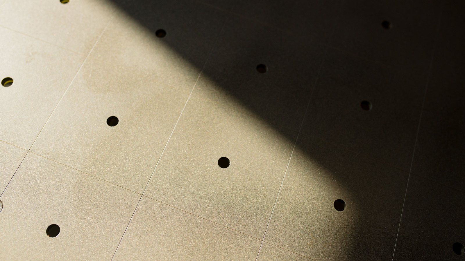 A pale, finely speckled flat surface photographed at an angle and filling the frame, drilled with round holes set in a wide regular grid and divided by faint straight seams. A hard-edged shadow cuts diagonally across the upper right, leaving that part of the surface almost black while the lower left stays brightly lit. No text, sign, object or person is in the picture.