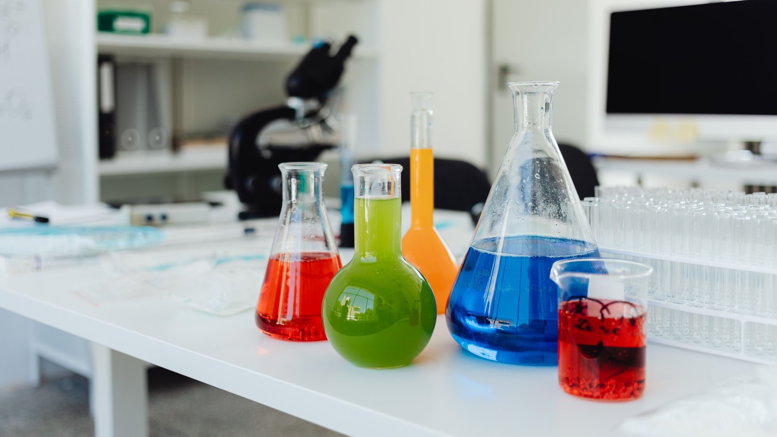 Glass flasks with colored liquids stand on an empty laboratory bench beside a microscope and test-tube racks.
