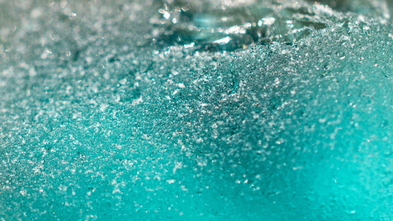 A close photograph of the frosted edge of a piece of teal glass, the surface broken into thousands of fine glittering facets that catch the light along the top of the frame and smooth into flat turquoise toward the bottom. The frame is filled edge to edge and holds no object, person, lettering or brand mark.