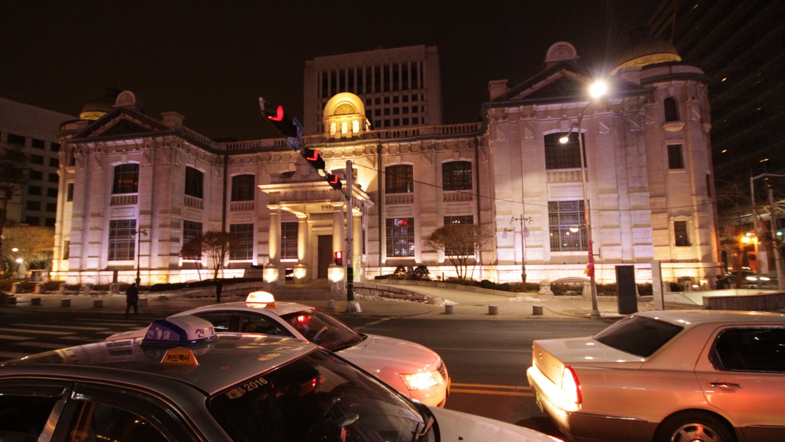 The Bank of Korea head office in Seoul floodlit at night, a stone building with a pedimented entrance and domed corner pavilions, taxis waiting at the crossing in front of it.