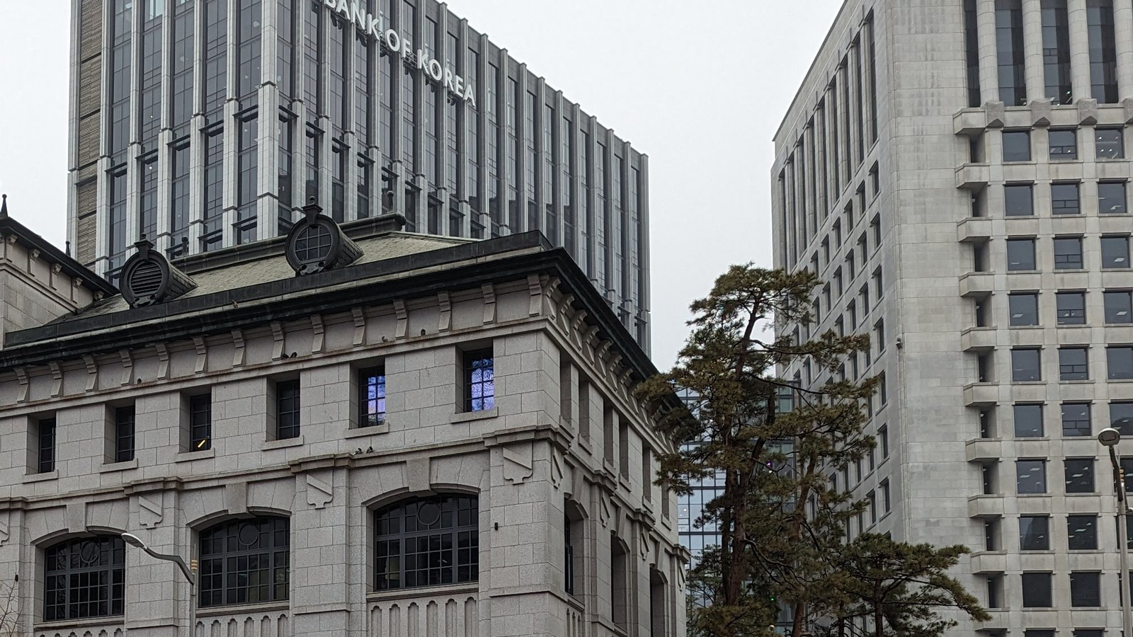 The raised lettering BANK OF KOREA across the upper floors of the central bank's grey office tower in Seoul, seen above the stone parapet, dormer roundels and arched windows of the older granite building in front of it, under flat white overcast, with a pine and a further stone office block to the right.