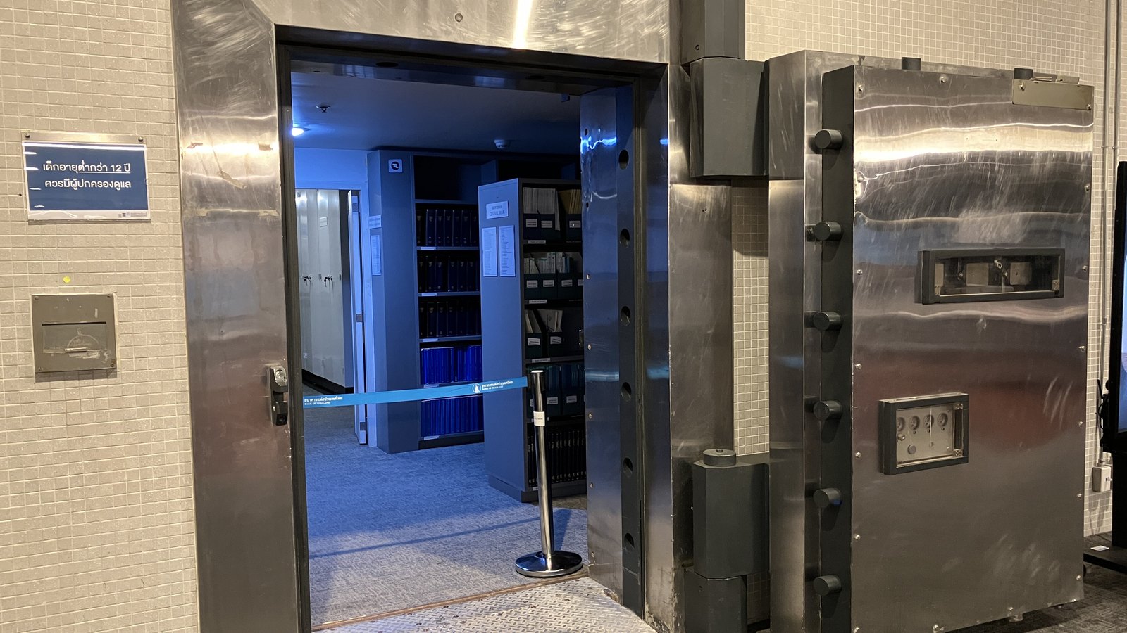 The open steel door of a strong room at the Bank of Thailand Learning Center in Bangkok, its edge studded with cylindrical locking bolts and its face carrying a small dial plate and a barred inspection panel. Through the doorway a blue lit vault holds shelves of archive boxes and ring binders, with a retractable barrier tape across the threshold printed with the Bank of Thailand name in Thai and English.