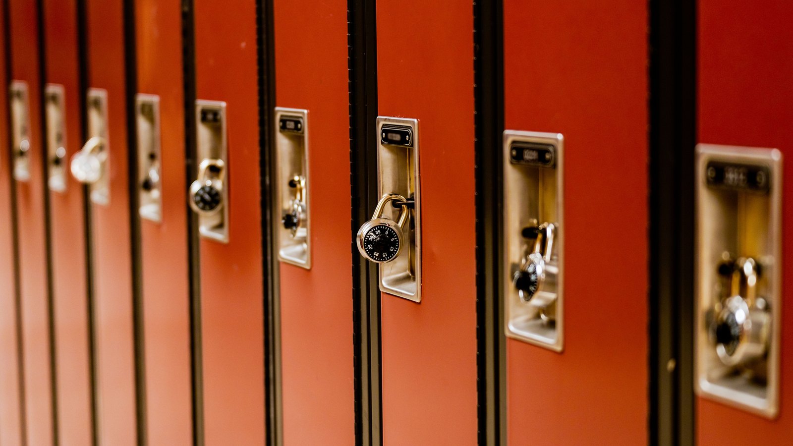 A close, shallow-focus crop of a row of red-orange school corridor lockers, each with a recessed metal handle and a black-dialled combination padlock, one padlock sharp in the centre of the frame and the rest falling away out of focus to the left and right. No person, no signage, no room number, no street and no landmark is visible. Stock photograph, not a photograph of this event.