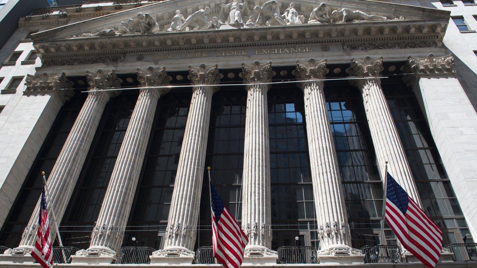The New York Stock Exchange seen from below, its Corinthian columns and sculpted pediment rising above the words New York Stock Exchange in gold lettering, with three United States flags hanging from the front.