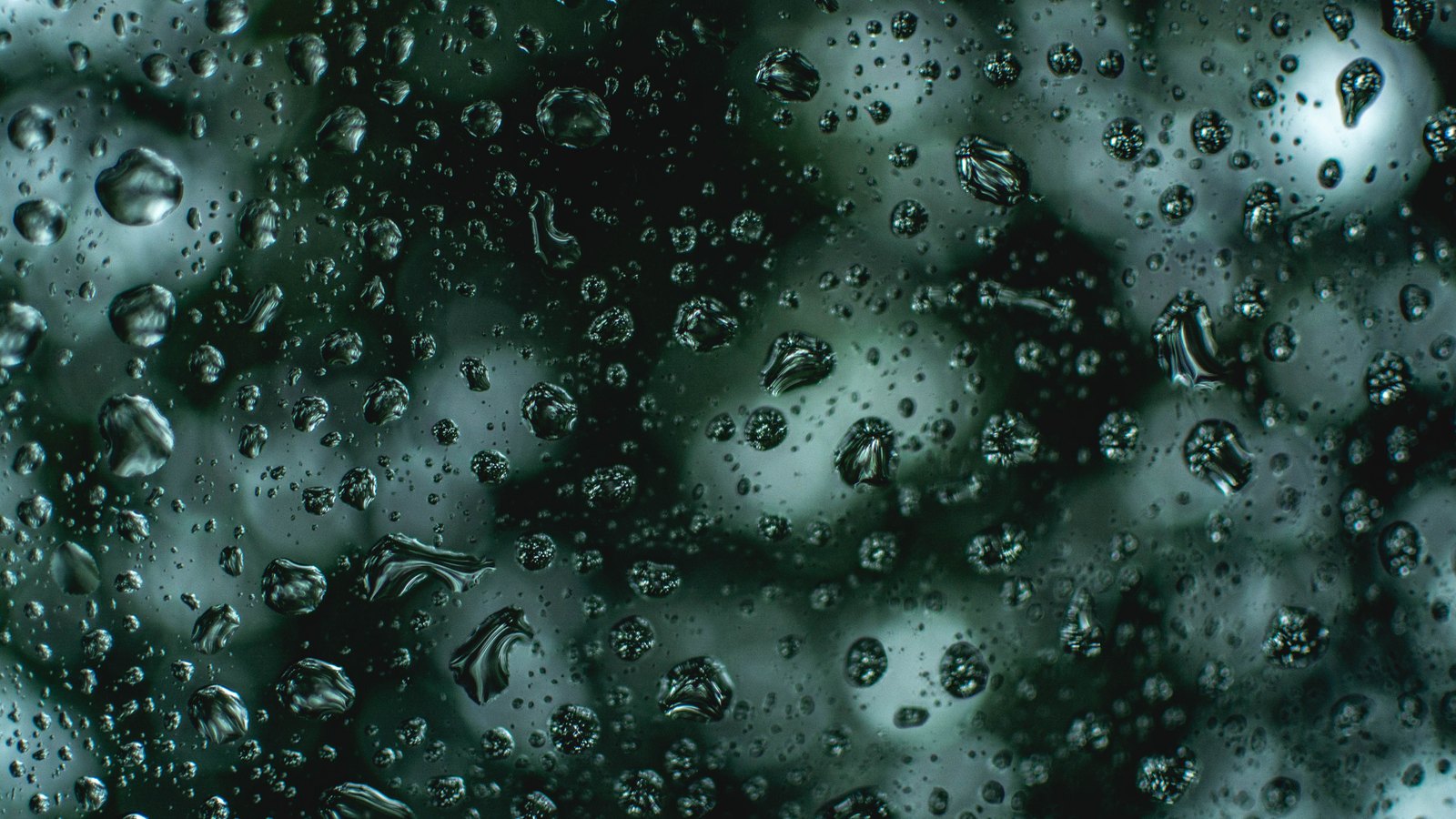 Macro of rain beaded on a pane of glass, the droplets catching a cold green light against near black behind them.