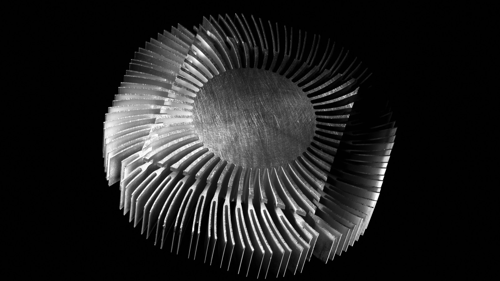 A radial aluminium heat sink photographed against a plain black background, its machined fins fanning out in a tight spiral from a brushed circular hub at the centre, lit from the left so the metal edges catch the light. No text, brand mark, person or place is in view.