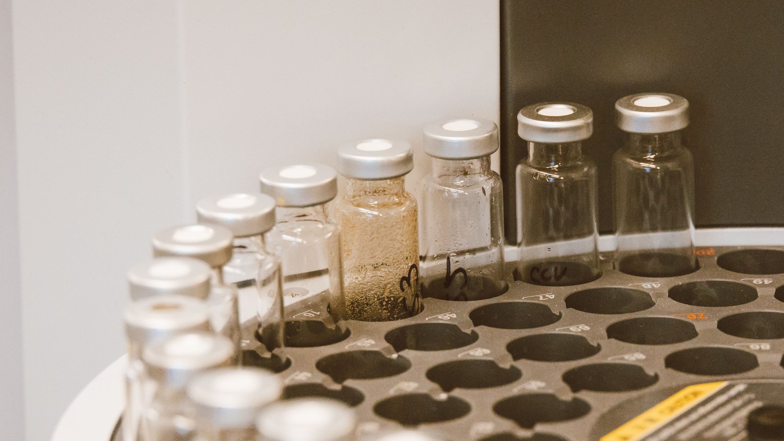 Sealed glass vials with crimped silver caps standing in the numbered wells of a laboratory autosampler carousel, one of them holding a cloudy amber liquid.