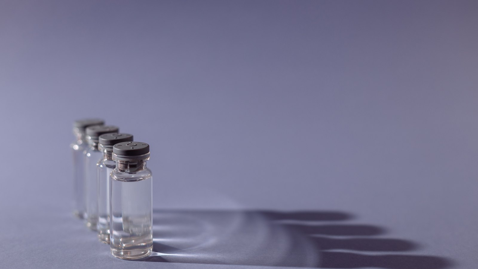 Four small clear glass vials with grey crimped stoppers standing in a diagonal row at the left of the frame on a plain pale grey surface, each holding a shallow layer of clear liquid, their long soft shadows falling away to the right across otherwise empty ground. No label or marking appears on any of them.