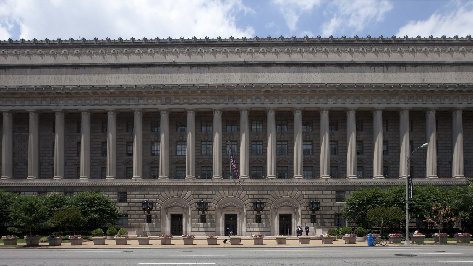 The long colonnaded front of the Herbert C. Hoover Building in Washington, headquarters of the United States Department of Commerce, with an inscription carved along the entablature, a United States flag on a pole at the centre and three arched doorways at street level.