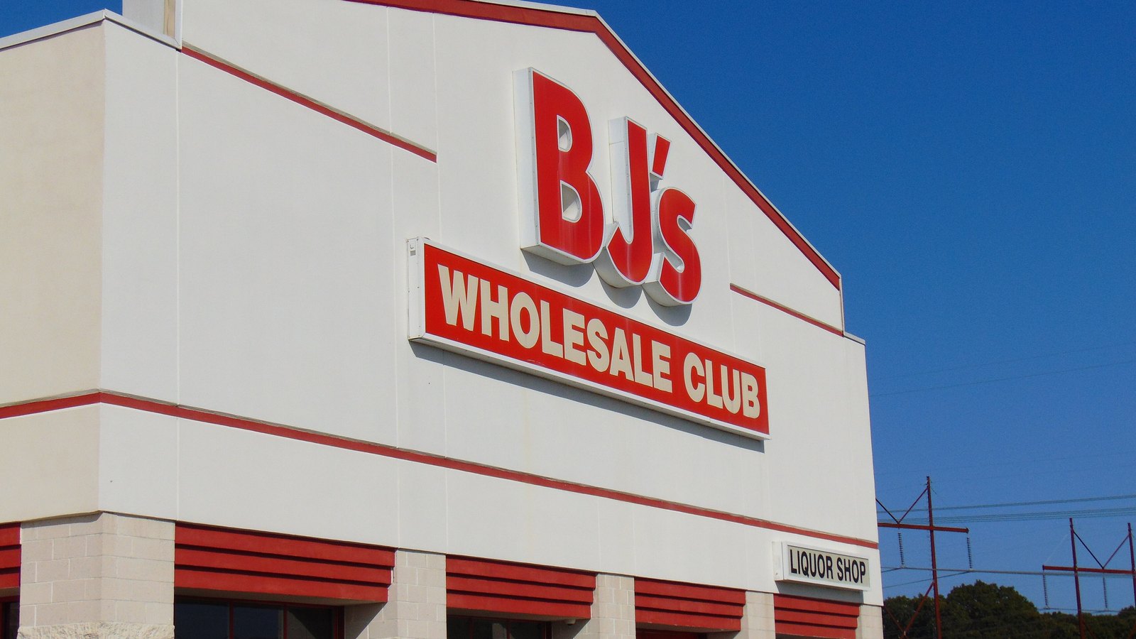 The corner of a BJ's Wholesale Club warehouse store, white cladding with red banding, the company name in raised red letters above a red sign panel and a smaller Liquor Shop sign at the right, under a clear blue sky. No person or vehicle is in the frame.