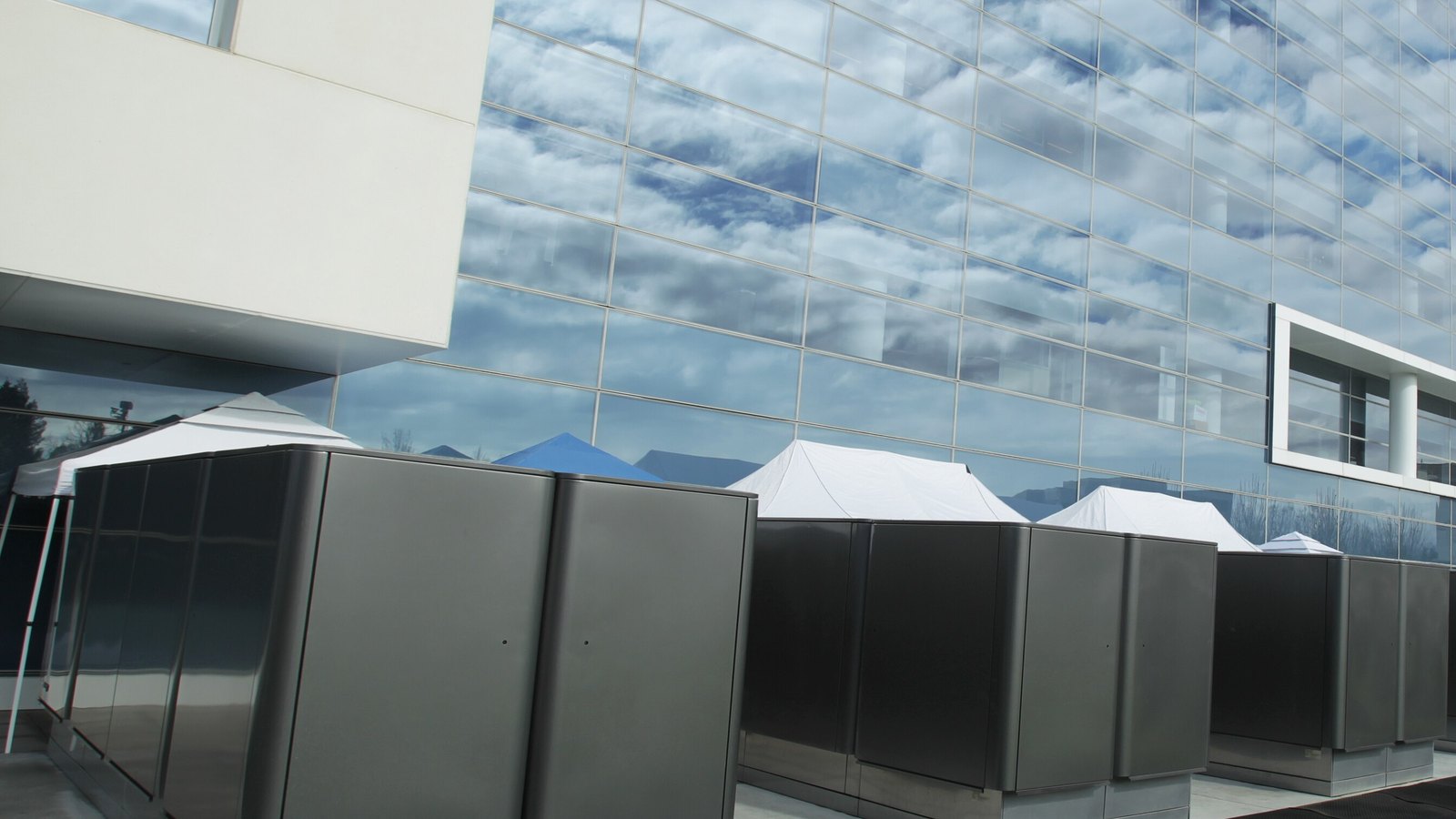 A row of grey Bloom Energy Server cabinets with white canopies above them, standing on paving beside a mirrored glass office facade that reflects white clouds and blue sky.