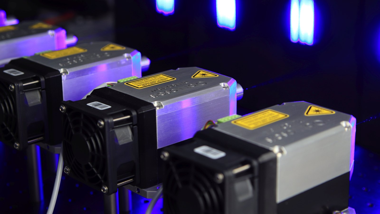 Four compact laser modules cast blue and violet light across an optics workbench